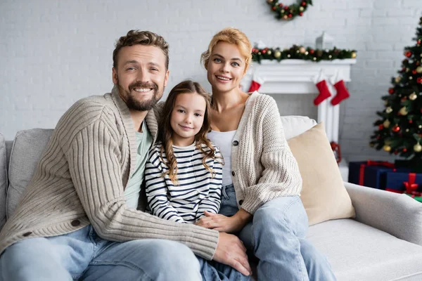 Fröhliche Familie blickt in die Kamera, während sie auf der Couch neben verschwommenem Weihnachtsdekor sitzt — Stockfoto