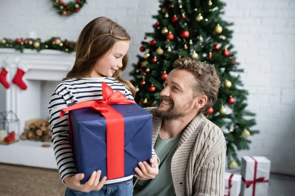 Sorrindo pai olhando para a filha com presente perto de árvore de Natal borrada em casa — Fotografia de Stock