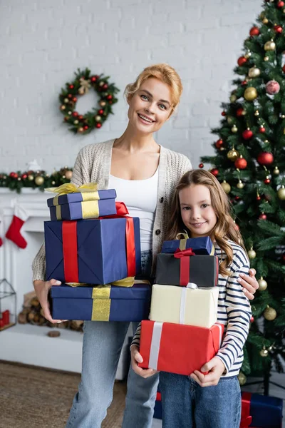 Sorrindo mãe e criança escondendo presentes e olhando para a câmera perto da árvore de natal em casa — Fotografia de Stock