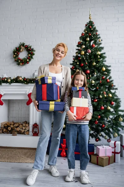 Cheerful mother and daughter holding presents near christmas tree and fireplace at home — Stock Photo
