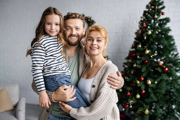 Sonriente hombre abrazando esposa e hija cerca borrosa árbol de Navidad en casa - foto de stock