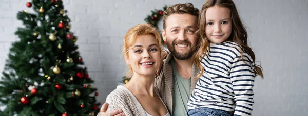 Positive man hugging child and wife near blurred decorated christmas tree at home, banner — Stock Photo