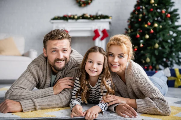 Famille positive regardant la caméra tout en étant allongé sur le tapis pendant la nouvelle année à la maison — Photo de stock