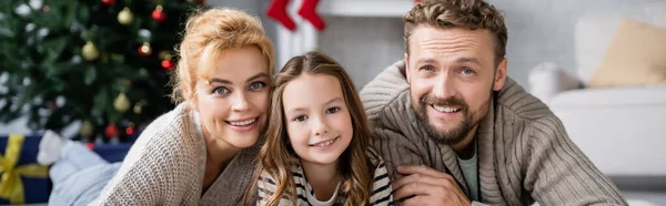 Cheerful parents looking at camera near daughter during new year celebration at home, banner — Stock Photo