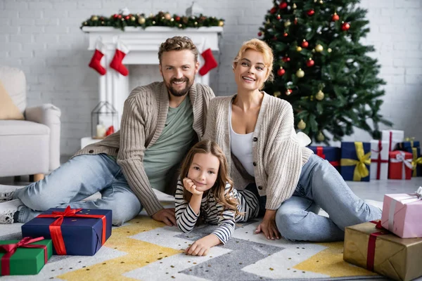 Lächelnde Familie blickt in die Kamera, während sie zu Hause neben Konfetti und Weihnachtsgeschenken auf dem Boden sitzt — Stockfoto