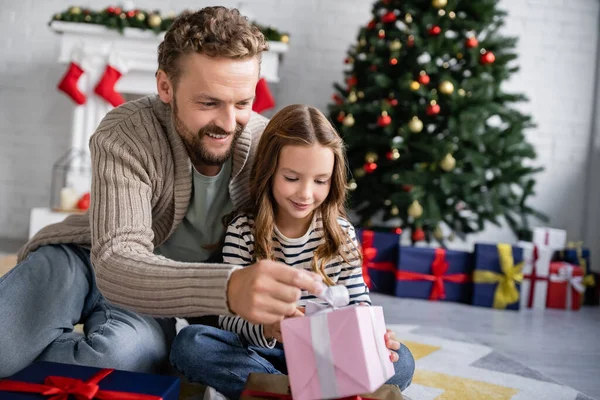 Uomo positivo guardando presente vicino figlia sul pavimento durante il Natale a casa — Foto stock