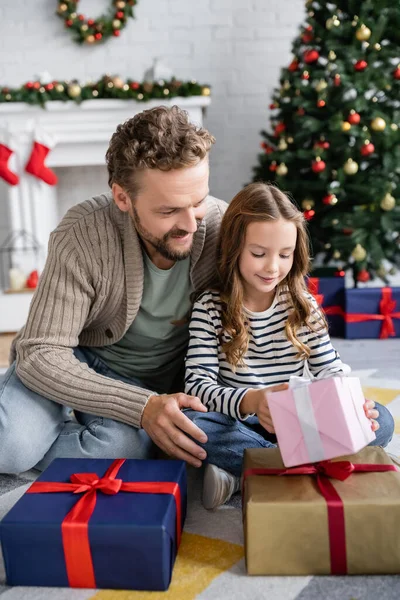 Lächelndes Mädchen hält Neujahrsgeschenk neben Papa auf Teppich im Wohnzimmer — Stockfoto