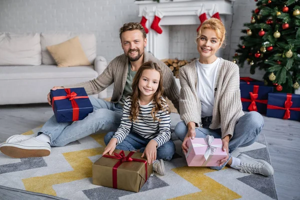 Positivo famiglia tenuta nuovo anno presenta e guardando la fotocamera sul tappeto a casa — Foto stock