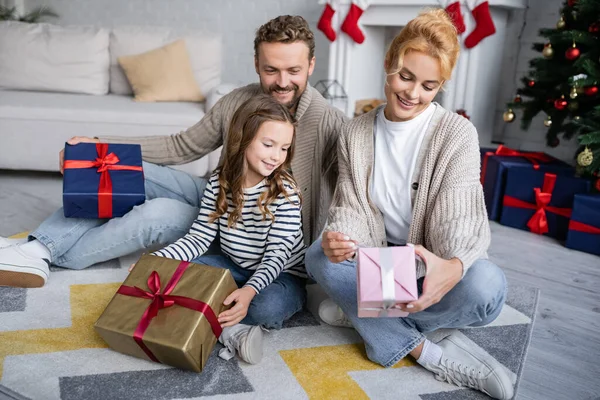 Mulher sorridente olhando para o presente de Natal perto da filha e do marido com presentes em casa — Stock Photo