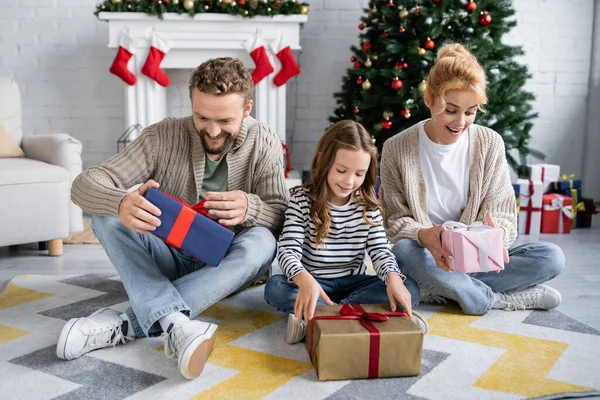 Fröhliche und aufgeregte Familie hält Weihnachtsgeschenke in der Hand, während sie auf Teppich im Wohnzimmer sitzt — Stockfoto