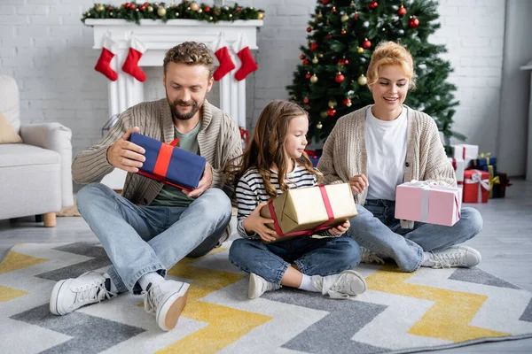 Niño sosteniendo regalo cerca de los padres sentados en el suelo en la sala de estar durante la celebración del año nuevo - foto de stock