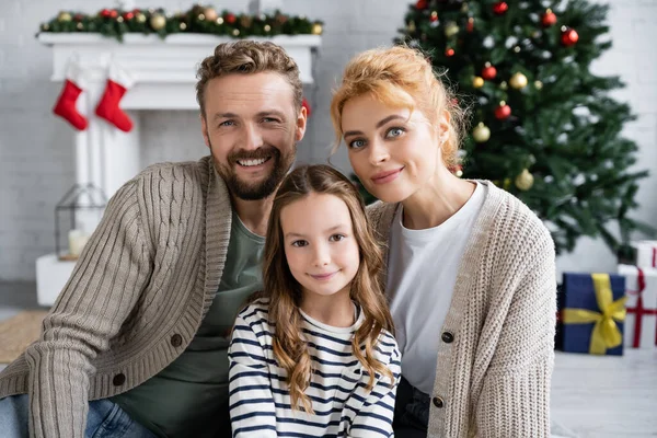 Famiglia sorridente con figlia che guarda la macchina fotografica vicino a decorazioni natalizie offuscate a casa — Foto stock