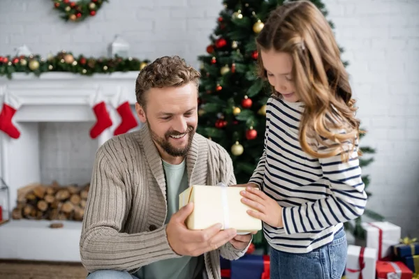 Homem positivo em malha cardigan segurando caixa de presente perto da criança durante o Natal em casa — Fotografia de Stock