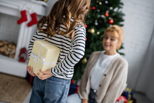 Chica ocultar regalo de Navidad cerca borrosa madre en casa - foto de stock