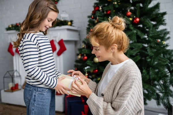 Side view of girl giving new year present to smiling mom in knitted cardigan at home — Stock Photo