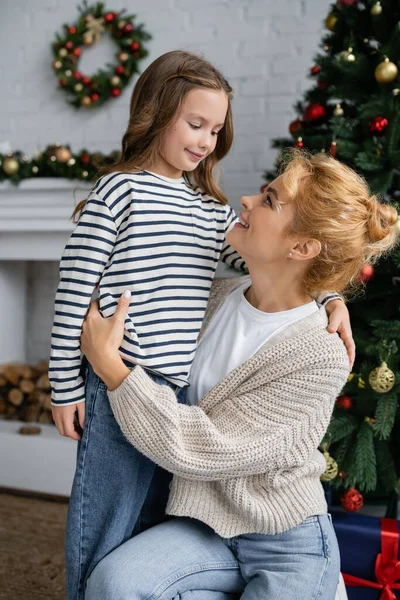 Mulher positiva em cardigan quente abraçando a filha sorridente durante a celebração do ano novo em casa — Fotografia de Stock