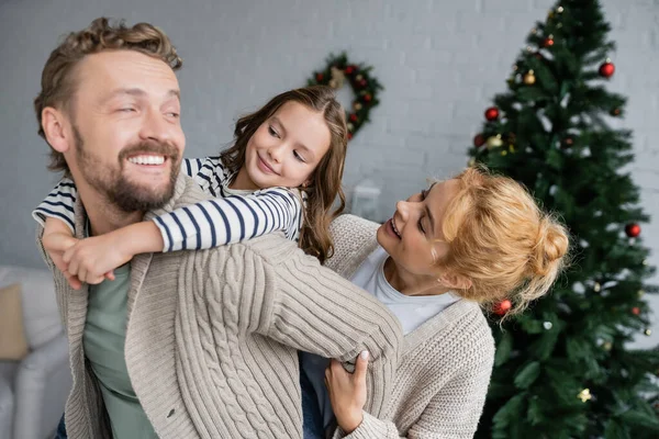 Chica alegre abrazando a papá cerca de la madre y borrosa árbol de Navidad en casa - foto de stock