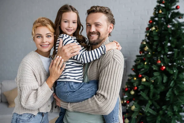 Père souriant dans un cardigan chaud tenant fille près de la femme lors de la célébration du nouvel an à la maison — Photo de stock