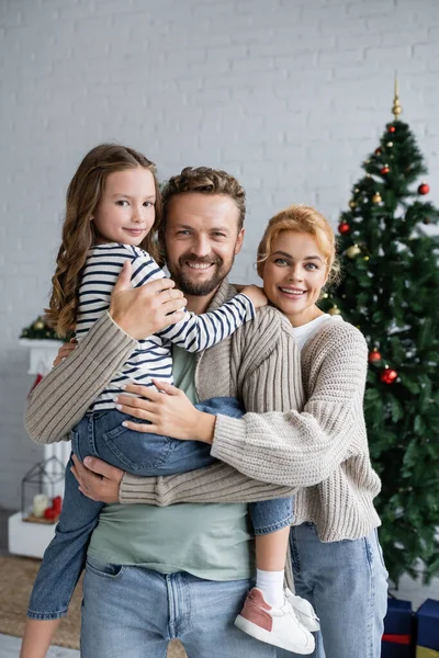 Позитивный мужчина держит дочь рядом с женой в кардигане и размытой рождественской елке дома — стоковое фото
