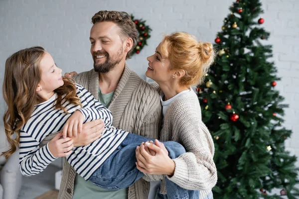 Genitori in cardigan che sorridono alla figlia vicino all'albero di Natale sfocato a casa — Foto stock