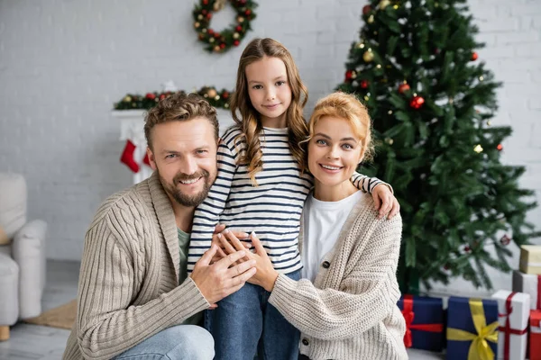 Alegre familia mirando a la cámara cerca de árbol de Navidad y borrosa regalos en casa - foto de stock