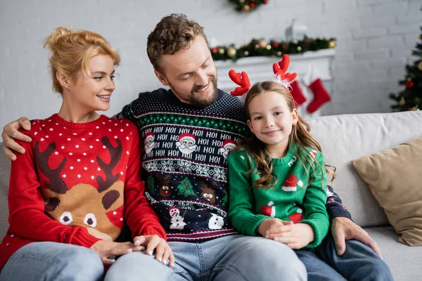 Homem alegre em camisola de Natal olhando para a criança e abraçando a esposa no sofá em casa — Fotografia de Stock