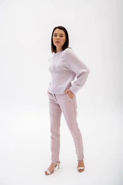 Full length of trendy brunette woman in knitted sweater and pants posing with hand in pocket on white background — Stock Photo