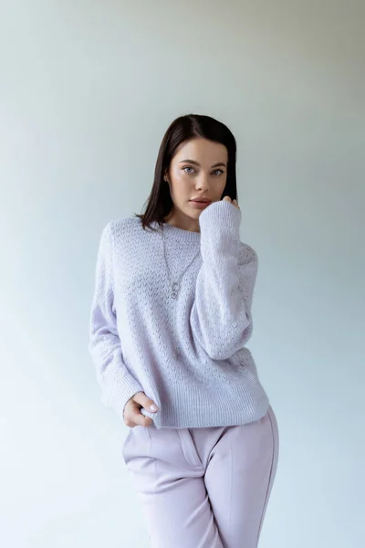 Pretty brunette woman in soft sweater holding hand near face and looking at camera isolated on grey — Stock Photo