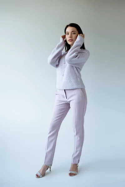 Full length of young woman in sweater and pants fixing hair while posing on grey background — Stock Photo