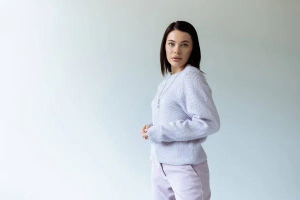 Brunette woman in knitted sweater and trousers looking at camera on grey background — Stock Photo