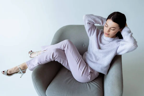 Full length of brunette woman in stylish clothes and sandals relaxing in armchair with closed eyes on grey background — Stock Photo