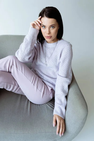 Donna bruna in maglione caldo e pantaloni seduti su una comoda poltrona isolata sul grigio — Foto stock