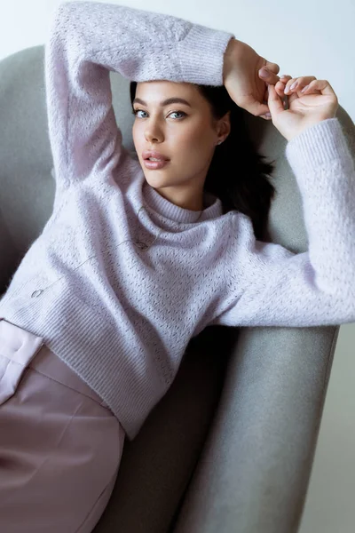 Vista ad alto angolo di giovane donna in maglia maglione seduta in poltrona con le mani sopra la testa isolata su grigio — Foto stock
