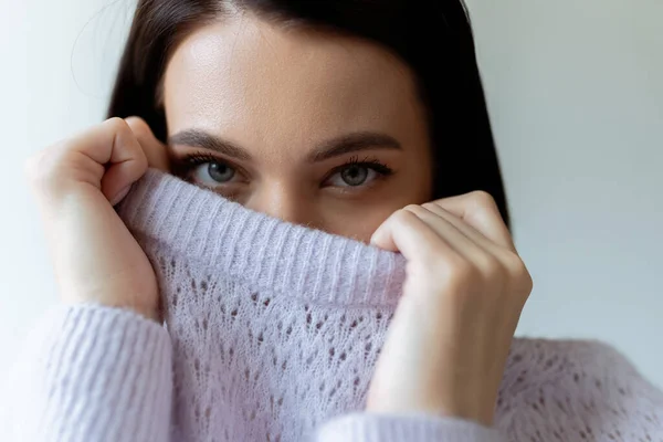 Young brunette woman looking at camera while obscuring face with soft knitted sweater isolated on grey — Stock Photo