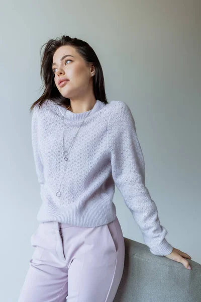 Mujer joven y elegante en suéter acogedor apoyado en el sillón y mirando hacia otro lado aislado en gris - foto de stock