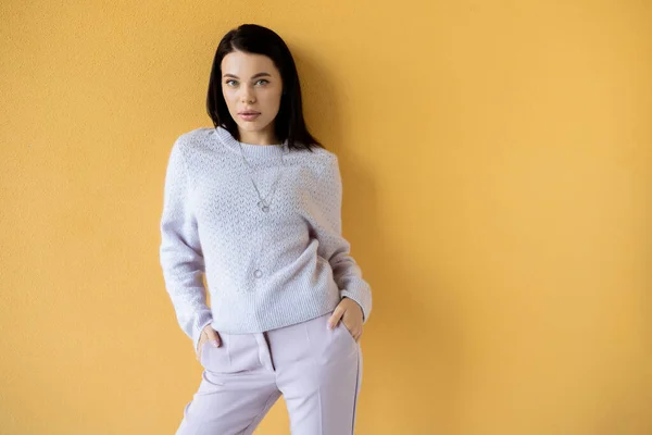 Brunette woman in soft knitted sweater standing with hands in pockets of trousers on yellow background — Stock Photo