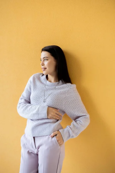 Mujer morena sonriente en suéter de punto posando con la mano en el bolsillo y mirando hacia otro lado sobre fondo amarillo - foto de stock