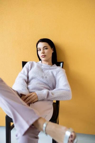 Модная женщина в уютном свитере смотрит в камеру, сидя на желтом фоне — стоковое фото