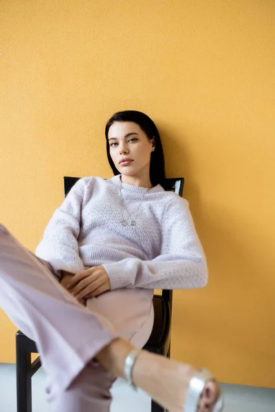 Mulher na moda em camisola acolhedora olhando para a câmera enquanto sentado no fundo amarelo — Fotografia de Stock