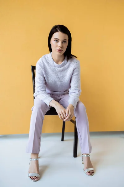 Full length of woman in knitted sweater and sandals sitting on chair on yellow background — Stock Photo