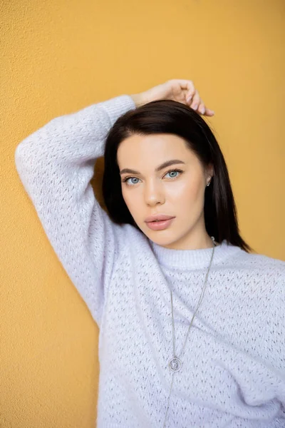 Young brunette woman in soft sweater posing with hand above head on yellow background — Stock Photo