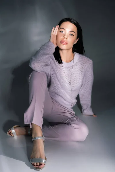 Volle Länge der brünetten Frau in trendigen Kleidern und Sandalen sitzt auf grauem Hintergrund mit Beleuchtung — Stockfoto