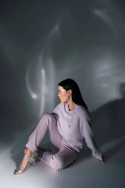 Full length of brunette woman in cozy sweater and sandals sitting on grey background with lighting — Stock Photo