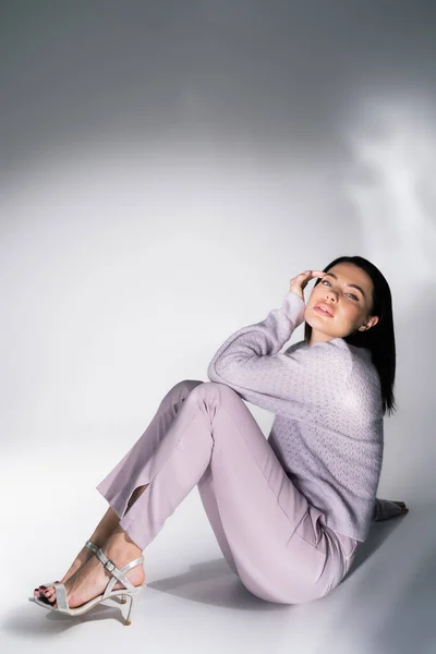 Charmante femme en pull tricoté et pantalon assis et regardant la caméra sur fond blanc et gris — Photo de stock