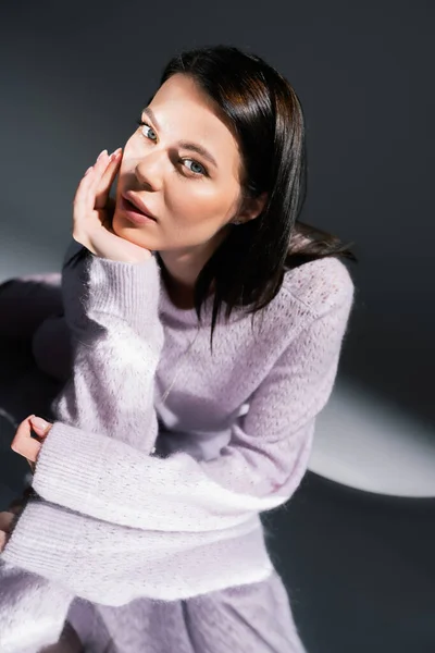 Sinnliche brünette Frau im warmen Pullover blickt in die Kamera, während sie auf grauem Hintergrund mit Beleuchtung liegt — Stockfoto