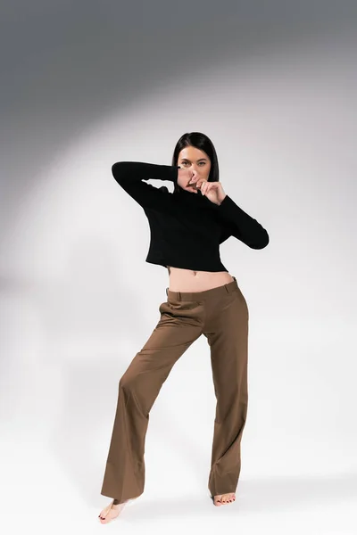 Full length of barefoot woman in brown pants and black sweater posing with hands near face on grey background — Stock Photo