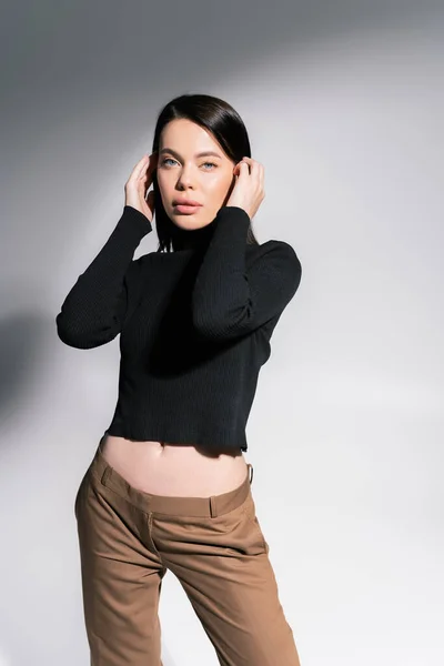 Mulher morena bonita em suéter preto fixando o cabelo e olhando para a câmera em fundo cinza e branco — Fotografia de Stock