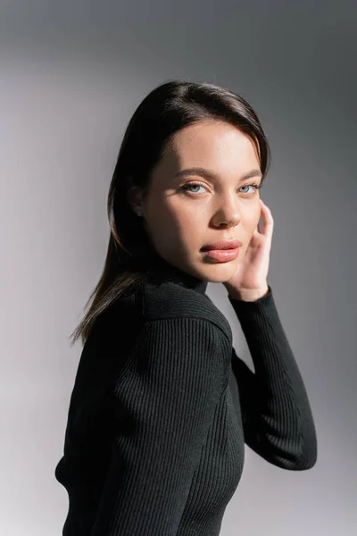 Brunette woman with natural makeup holding hand near face and looking at camera on grey background — Stock Photo