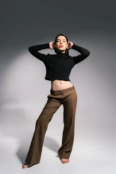Longitud completa de mujer descalza en suéter negro y pantalones marrones posando sobre fondo gris - foto de stock