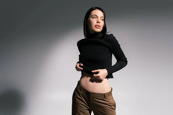 Sensuale donna bruna in maglione nero e pantaloni marroni guardando lontano su sfondo grigio — Foto stock